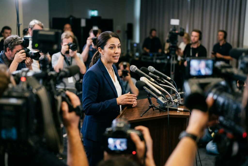 Spokesperson speaking at a press conference podium surrounded by microphones and TV cameras